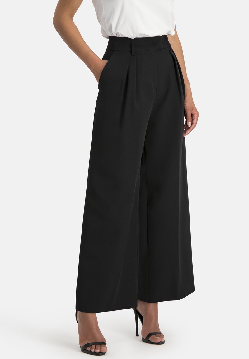 Wijde zwarte broek van soepel materiaal, met voorplooien, een hoge taille en zijzakken. Gecombineerd met zwarte haksandalen.