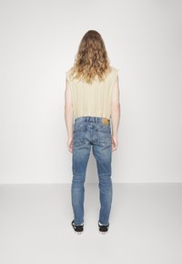Persona con cabello largo y rizado, vistiendo una camiseta sin mangas color beige, jeans azules y zapatillas negras, de pie frente a una pared blanca lisa.