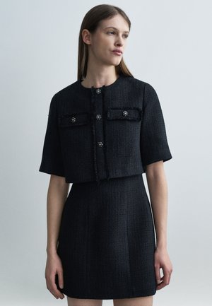 Mujer con chaqueta negra de manga corta con textura y botones decorativos, y falda a juego de talle alto, posando frente a un fondo liso.