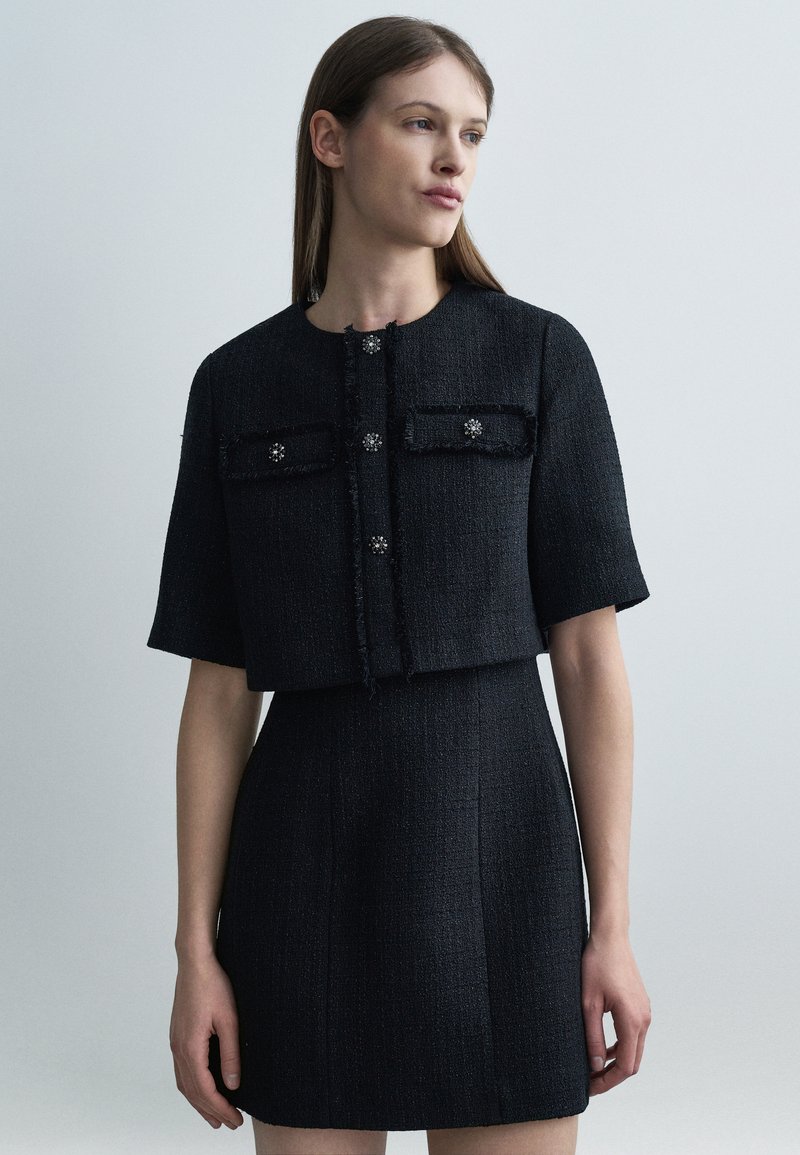 Γυναίκα που φοράει μαύρο υφασμάτινο σακάκι με κοντά μανίκια και διακοσμητικά κουμπιά, μαζί με ταιριαστή ψηλόμεση φούστα, στέκεται μπροστά σε απλό φόντο.