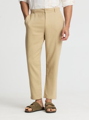 Uomo che indossa pantaloni beige a gamba dritta con bottone frontale e tasche, abbinati a sandali verde oliva con punta aperta.