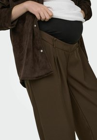 Brūns zamša krekls ar spraudņu pogām, pārklāts ar melnu pieguļošu topu un brūnām plīsētām biksēm, kas demonstrē atslābinātu un strukturētu dizainu.