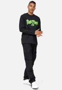 Sort langærmet skjorte med grønt grafisk logo, sammen med sorte cargo bukser og hvide sneakers, der har praktiske lommer og et stilrent design.