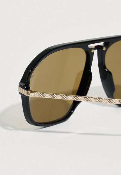 Occhiali da sole neri con lenti marroni. La montatura è lucida con una forma quadrata. Le aste presentano un dettaglio dorato testurizzato vicino alla cerniera.