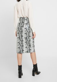 Kvinna iklädd en hög midja, knälång kjol med ormskinnsmönster och bakdragkedja, krämfärgad blus med spetsärmar och svarta klackiga ankelstövlar.