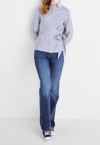 Chemise rayée à boutons avec une taille nouée en bleu et blanc, associée à un jean bleu évasé et des baskets en toile marine.