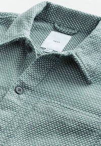 Green textured shirt with a checkered pattern, featuring a fold-down collar and two dark buttons on the front. Label reads "NEXT."