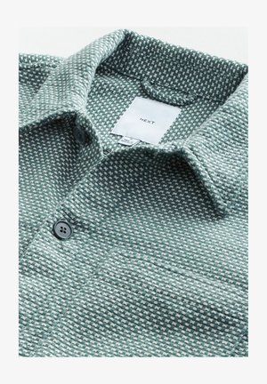 Grünes, strukturiertes Hemd mit karierter Musterung, ausgestattet mit einem umklappbaren Kragen und zwei dunklen Knöpfen auf der Vorderseite. Das Etikett trägt die Aufschrift "NEXT".