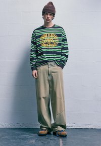 Dlhý rukáv pruhované tričko v zelenej a čiernej farbe so žltou grafikou, kombinované s voľnými béžovými nohavicami a hnedými štruktúrovanými topánkami s modrými šnúrkami.