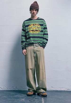 Dlhý rukáv pruhované tričko v zelenej a čiernej farbe so žltou grafikou, kombinované s voľnými béžovými nohavicami a hnedými štruktúrovanými topánkami s modrými šnúrkami.