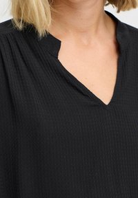 Kvinna i en svart texturerad blus med en nedskuren V-ringning och kort blont hår synligt vid kragen.