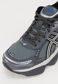 Gros plan sur une chaussure de course Asics grise et noire avec tissu en maille, accents réfléchissants et lacets noirs.