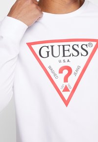 Persona vistiendo una camiseta de manga larga blanca con un logo de triángulo rojo que dice "GUESS U.S.A." y "WASHED JEANS" alrededor de un gran signo de interrogación rojo.