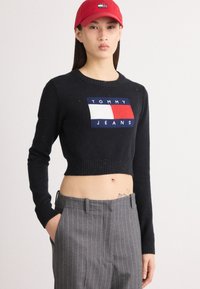 Černý zkrácený svetr s žebrovaným lemem, s výšivkou "TOMMY JEANS" v červené, bílé a modré barvě. V kombinaci se šedými kalhotami s jemným proužkem. Nosit červenou čepici.