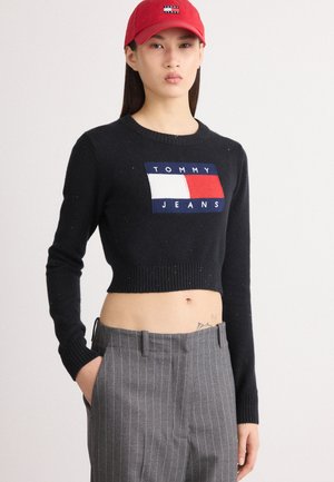 Maglione nero corto con orlo a coste, caratterizzato dal logo "TOMMY JEANS" rosso, bianco e blu. Abbinato a pantaloni grigi a righe. Cappellino rosso indossato.