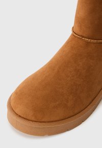 Detailní záběr hnědé zimní boty z nubukové kůže se šitými detaily a texturovanou gumovou podrážkou na bílém pozadí.