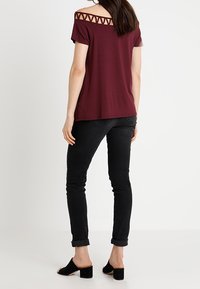 Burgundy off-shoulder topp med triangulär utskärning vid halsringningen, tillsammans med svarta åtsittande jeans och blockklackade mules.