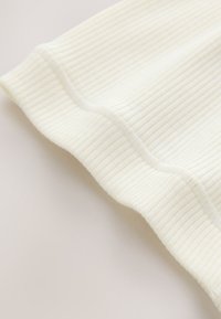 Gros plan d'un coin de tissu tricoté côtelé blanc avec un détail de couture visible sur un fond clair.