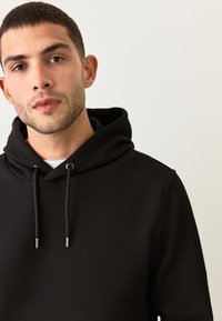 Felpa nera con cappuccio e motivo testurizzato. Presenta coulisse con punte in metallo. Design aderente, cuciture visibili e tessuto morbido.