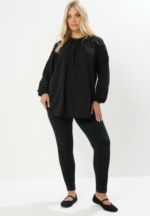Schwarzes langärmliges Oberteil mit gerafftem Ausschnitt und Spitzenakzenten an den Schultern, kombiniert mit schwarzen Slim-Fit-Hosen und schwarzen flachen Schuhen.