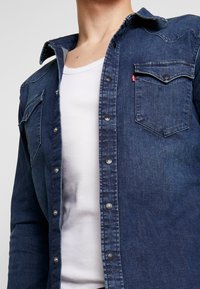 Mörkblå denimskjorta med knäppning, två bröstfickor och spetsig krage. Har en blekt textur och metallknappar.