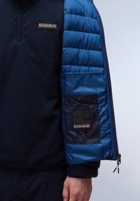 Sudadera de forro polar azul marino con mangas y bolsillo acolchados en azul; presenta detalles de logo y cierre de cremallera; textura suave y colores en contraste.