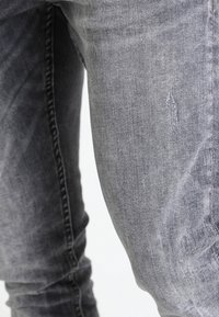 Vaqueros de mezclilla gris con un acabado deslavado, que presentan un ligero detalle desgastado en el muslo. Ajustados al cuerpo con costuras visibles.