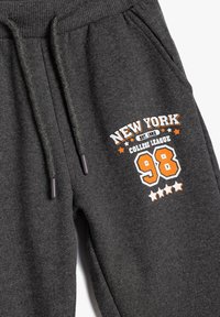 Pantalones de chándal grises con cintura elástica, cordón y bolsillos laterales. Presenta la impresión "New York College League" y el número "98" en naranja y blanco.