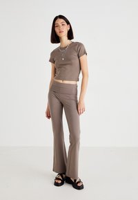 Mlada žena stoji, odjevena u taupe uski crop top, usklađene flare hlače, slojevite srebrne ogrlice i crne platforme na bijeloj pozadini.