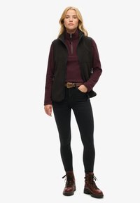 Czarna polarowa kamizelka na burgundowym golfie, połączona z czarnymi dopasowanymi dżinsami i brązowymi butami na sznurówki. Zawiera zamki błyskawiczne i brązowy pasek.