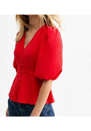 Donna che indossa una camicetta rossa brillante con trama, maniche a sbuffo, bottoni sul davanti e scollo a V, abbinata a jeans blu scuro.