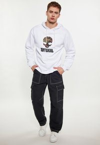 Fehér kapucnis pulóver katonai mintás koponya grafikával és fekete szöveggel "TUFFSKULL", fekete cargo nadrággal, fehér varrással és zsebekkel. Fehér sportcipő.
