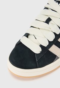 Black suede sneaker with beige stripes, featuring white laces, showcasing a close-up view of the toe and upper.