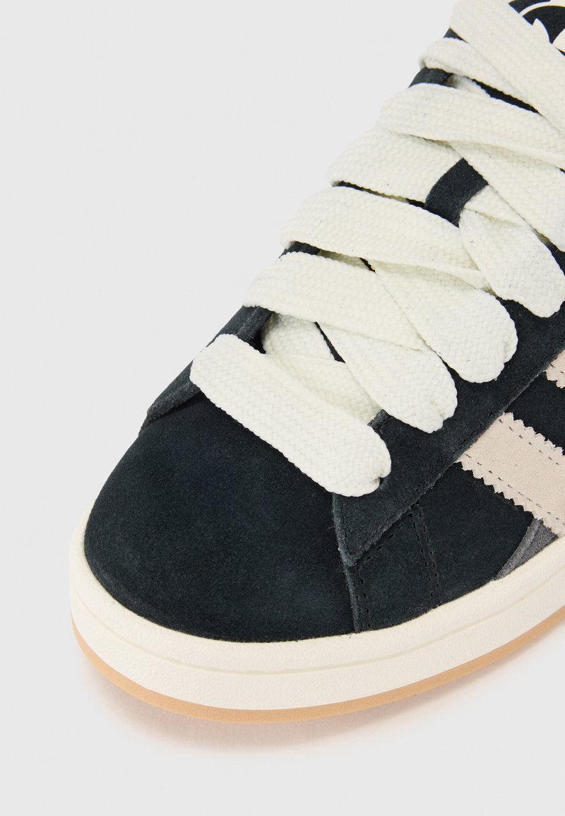 Zapatilla de ante negra con rayas beiges, con cordones blancos, mostrando una vista en primer plano de la punta y la parte superior.