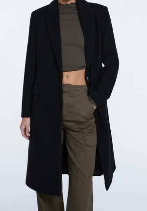 Kobieta w czarnym długim płaszczu, nałożonym na oliwkowozielony prążkowany krótki top i spodnie cargo z wysokim stanem na jednolitym białym tle.