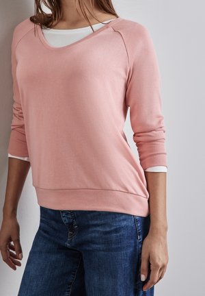 Femme portant un haut à manches longues rose clair superposé sur une chemise blanche et un jean bleu, debout devant un mur clair uni.