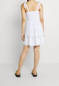 Žena v bílé šaty s vrstevnatým designem a zavázanými ramínky, s béžovými sandály, stojící na jednobarevném světlejším pozadí.