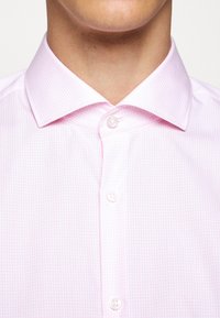 Gros plan d'une personne portant une chemise à carreaux rose clair avec un col ouvert boutonné en haut.