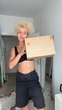 Braune Pappschachtel mit dem bedruckten Text "Love me. Wear me." in Weiß, gehalten von einer Person, die ein schwarzes Crop-Top und dunkle Shorts trägt.