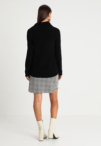 Pull noir avec un large col, associé à une jupe à carreaux grise. Le mannequin porte des bottines à talons carrés crème. Texture lisse et design ajusté.