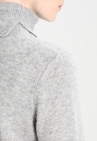 Hellgrauer gestrickter Pullover mit einem hohen, gerippten Kragen und einer weichen Textur, der ein tailliertes Design und subtile Nähdetails aufweist.