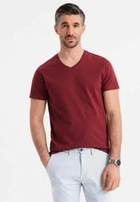 Camiseta de algodón de color burdeos con cuello en V, diseño ajustado. Mangas cortas, textura suave. Combinada con pantalones de color claro y un reloj.
