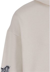 Lys beige hoodie med struktureret stof, ribstrikkede manchetter og et stort sort grafisk design på venstre ærme.