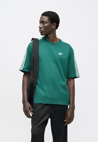 Zelené tričko s krátkym rukávom a bielymi tromi pruhmi na ramenách, s malým bielym logom na hrudi, v kombinácii s čiernymi nohavicami.