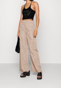 En person med lockigt hår bär en svart korttank topp och beige cargobyxor, håller en svart väska och står mot en grå bakgrund.