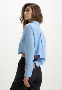 Camicia azzurra chiara cropped con maniche lunghe, polsini con bottoni, dettaglio con piega sul retro e un tessuto morbido e leggero. Abbinata a pantaloni neri.