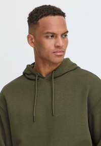 Olijfgroene hoodie met een trekkoord, zachte textuur en een relaxte pasvorm. Voorzien van een grote buidelzak en geribbelde manchetten.