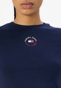 Tamsiai mėlyna medvilnės marškinėliai su apvalia grafika, kurioje užrašyta "tommy jeans" ir "new york", papuošti mažu raudonos ir baltos spalvos logotipu.