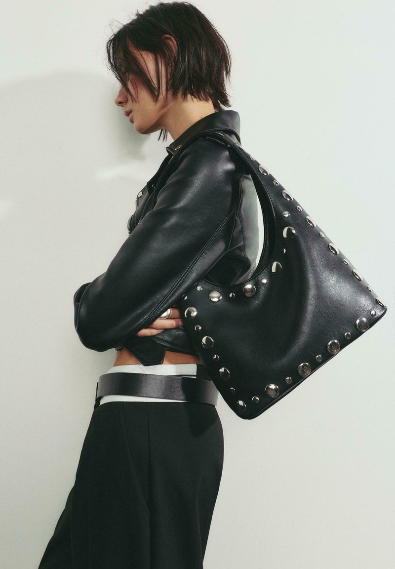 Czarna skórzana torba z srebrnymi ćwiekami, o zaokrąglonym kształcie i pojedynczym paskiem na ramię, w połączeniu z dopasowaną czarną skórzaną kurtką.