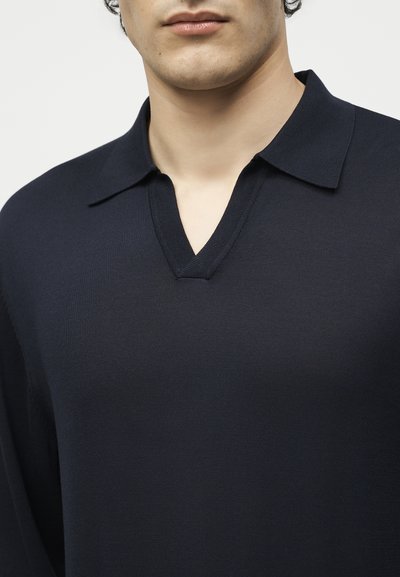 Άνδρας που φοράει σκούρο μπλε πλεκτό πουλόβερ με ανοικτό V λαιμόκοψη, εμφανίζοντας τον λαιμό και το κάτω μέρος του προσώπου σε ουδέτερο φόντο.